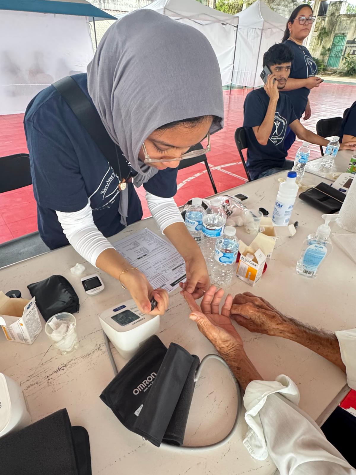 A young woman holds steady the hand of an elderly person who is out of the frame of the photo to prick her finger for a blood draw.