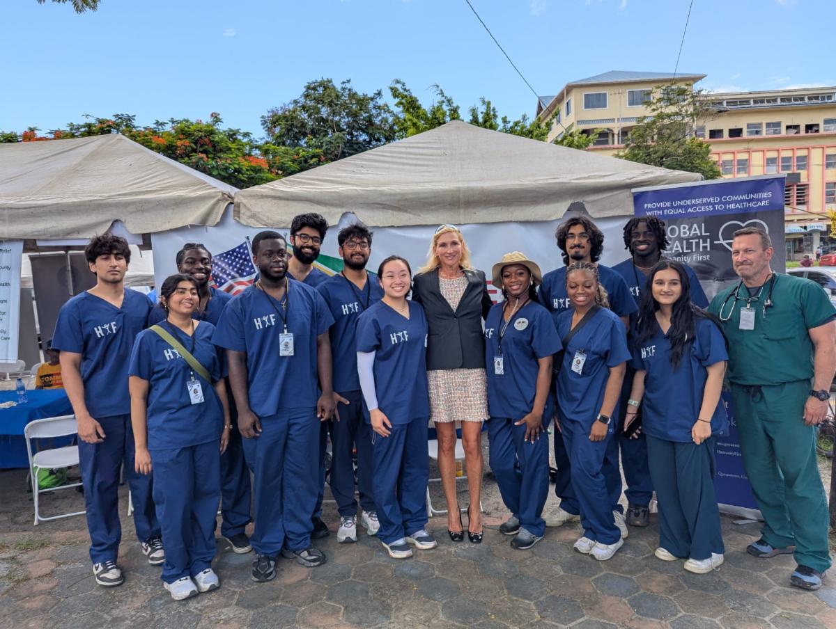 A group of young people wearing blue medical scrubs and one middle aged person wearing green scrubs pose with a woman in dress who is the US Ambassador to Guyana.