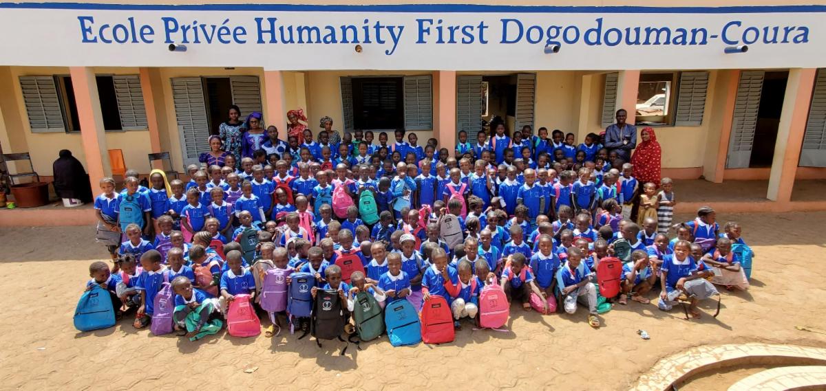 Several dozen primary school students pose with their teachers for a class photo outside the Humanity First school in Dogodomacoura Mali. They wear matching blue Humanity First smocks and hold their colorful backpacks.