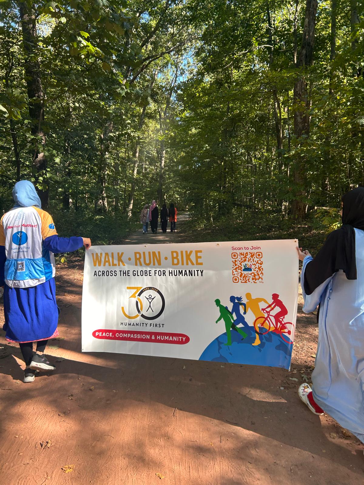 Two ladies in long clothes one of whom wears a humanity first cycling jersey over her clothes carry a large banner for the humanity first walk run bike event on a wide path through the woods