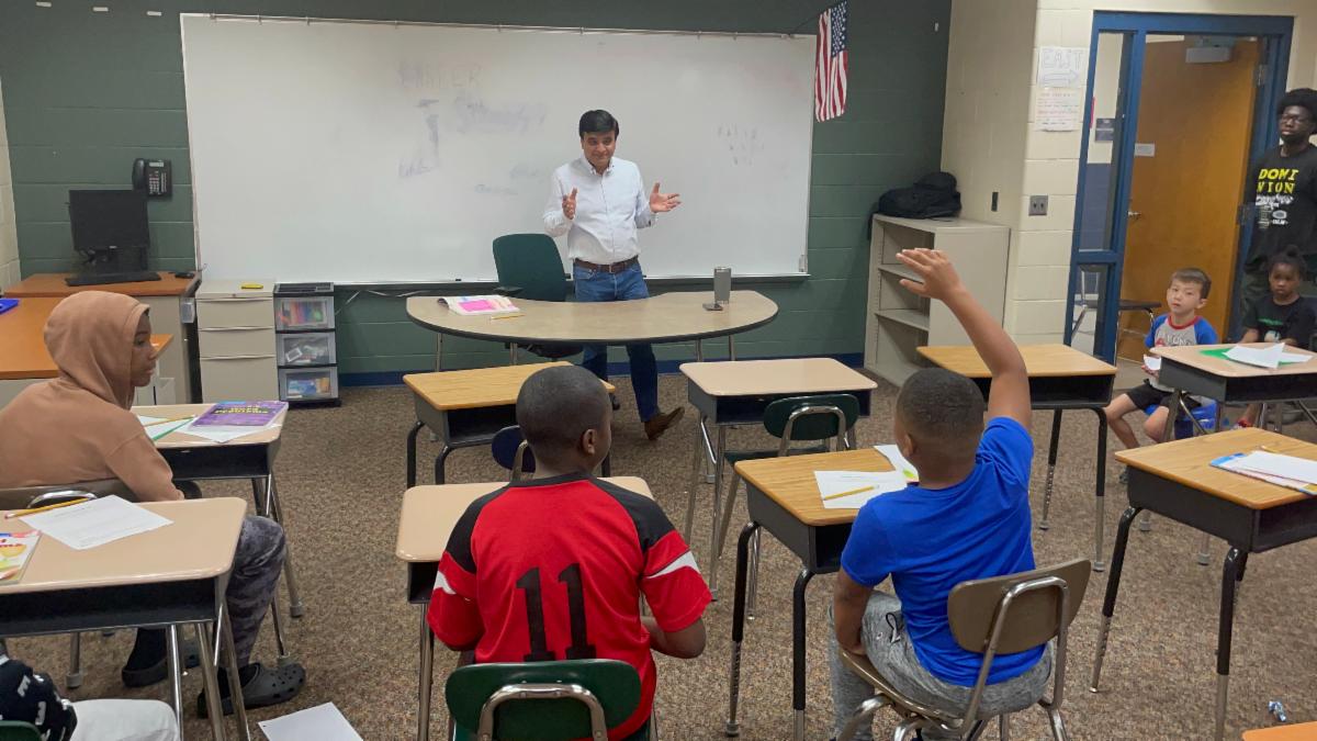 A man speaks animatedly to a group of seated students from the front of the classroom. One student has his hand raised to ask a question.