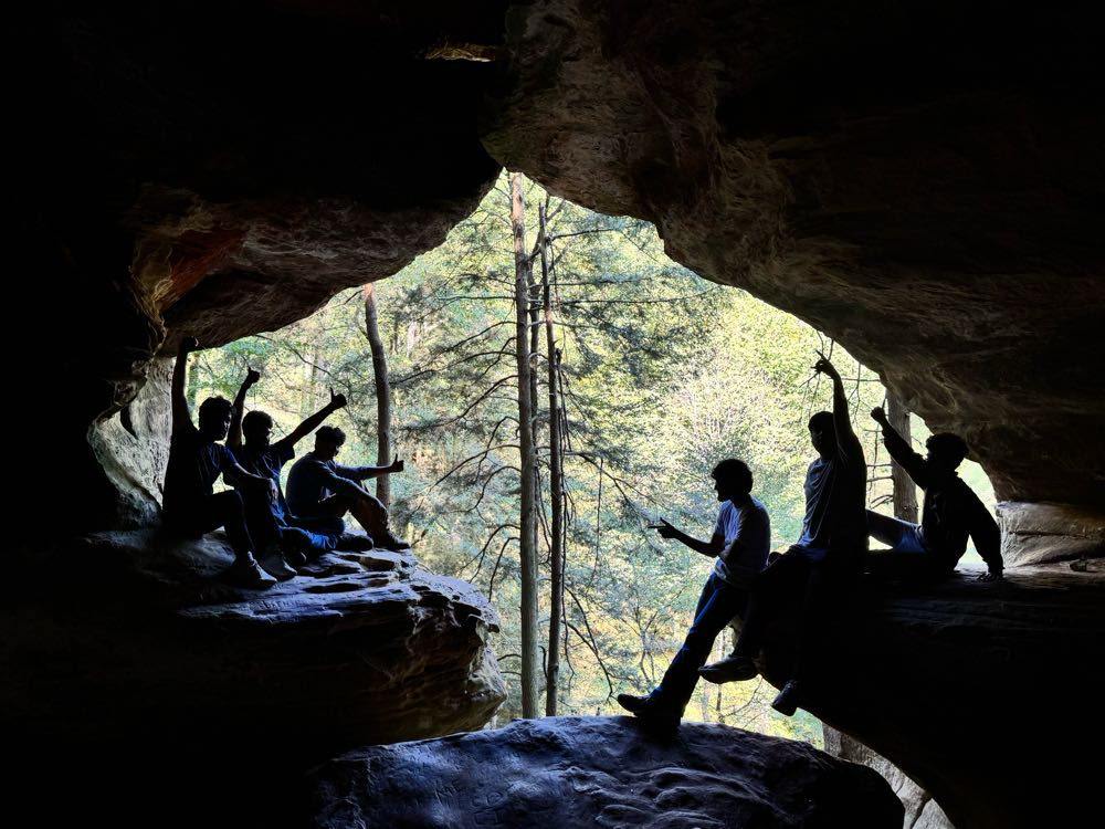 From inside a cave the photo shows the backlit silhouettes of six people with trees outside the opening of the cave.