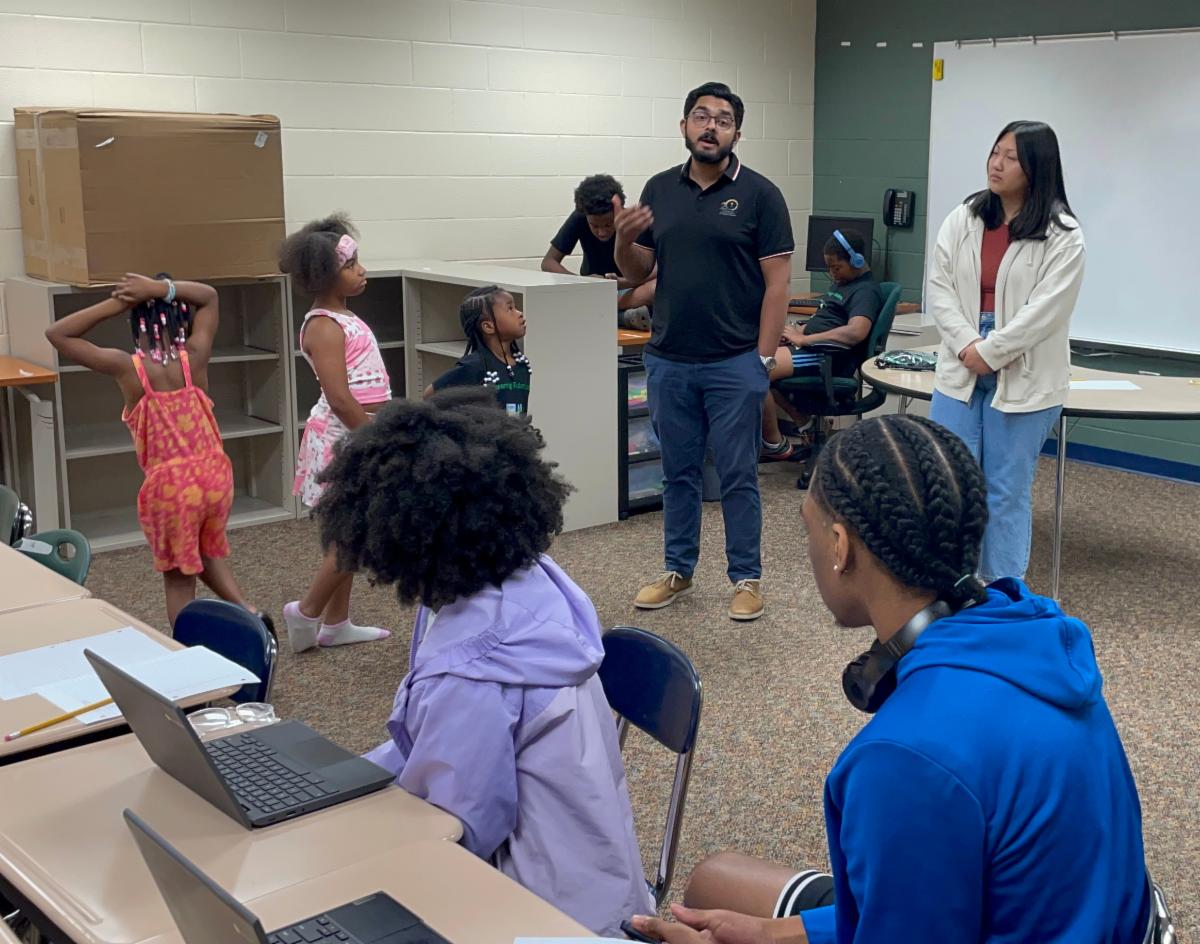 Two post-secondary students stand at the front of the classroom speaking to seated and standing students of varying ages. 