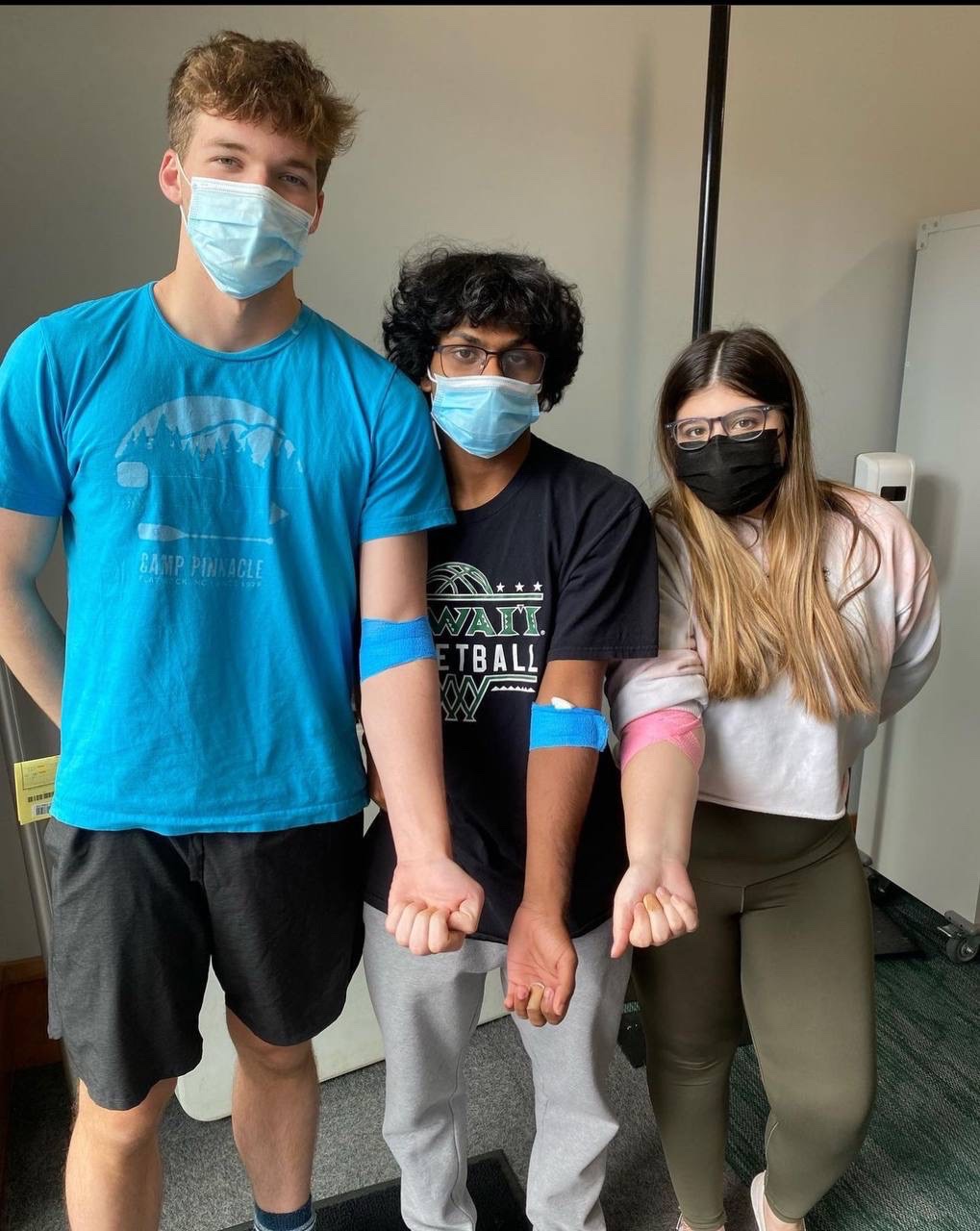 Three students stand side by side holding out their arms that have blue gauze at the elbow after getting blood drawn.