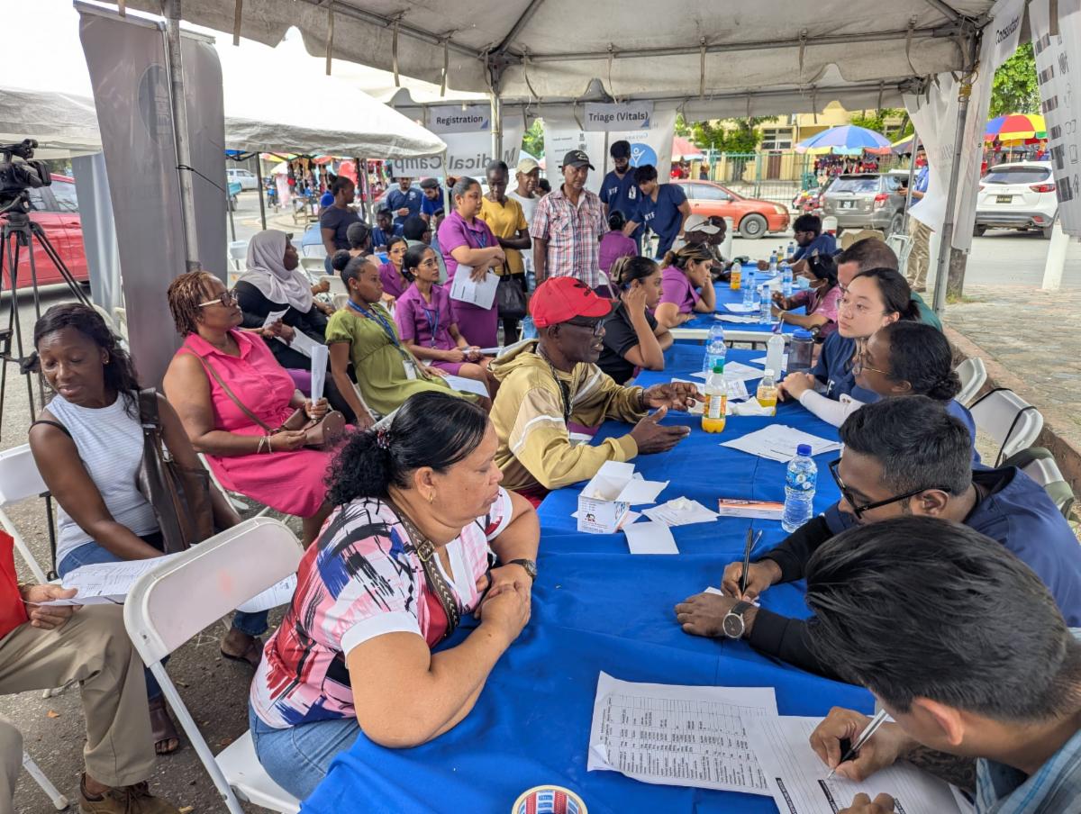 Under an open wall tent a long table with blue tablecloths is set up. Chairs on each side are filled with people in blue medical scrubs on one side and people in plain clothes on the other talking to each other. More people sit on an adjacent row of chairs