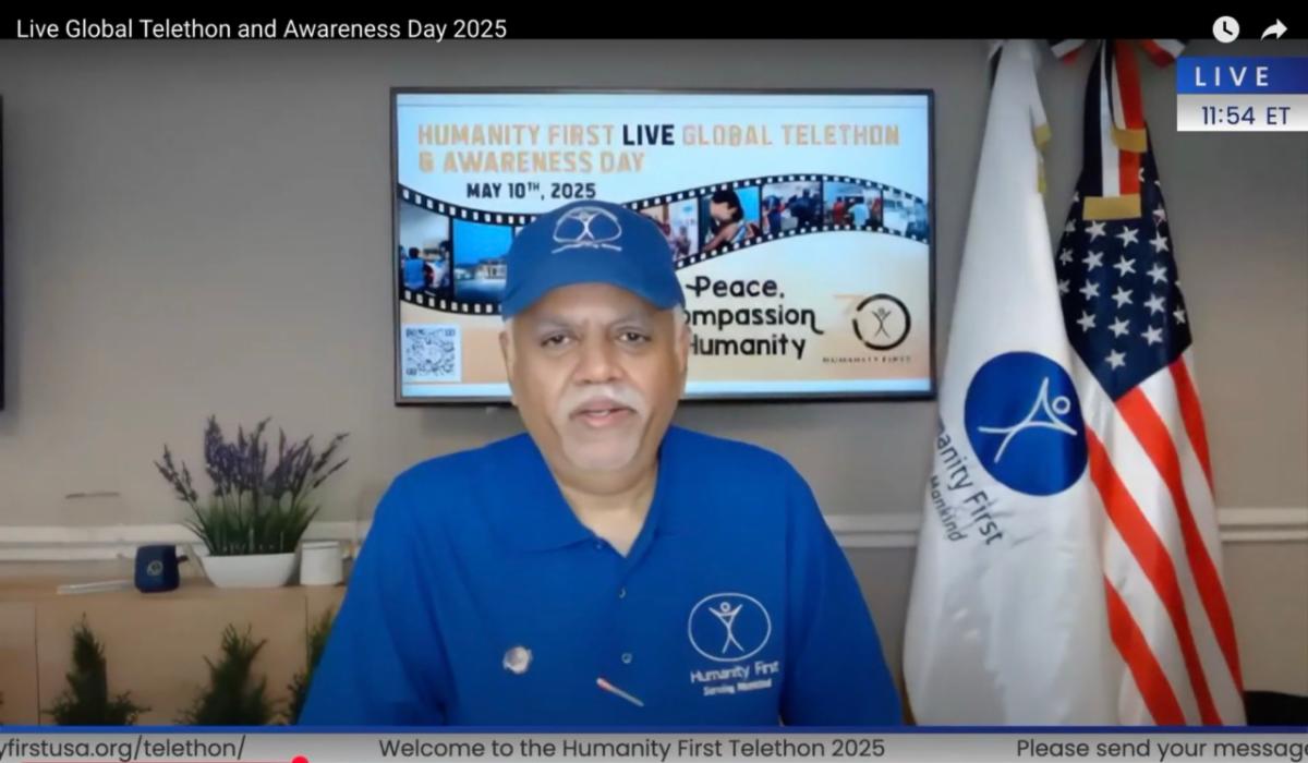 A man wearing a blue HF polo shirt and blue HF baseball cap sits in front of a digital design and two flags on stands representing Humanity First and the United States of America