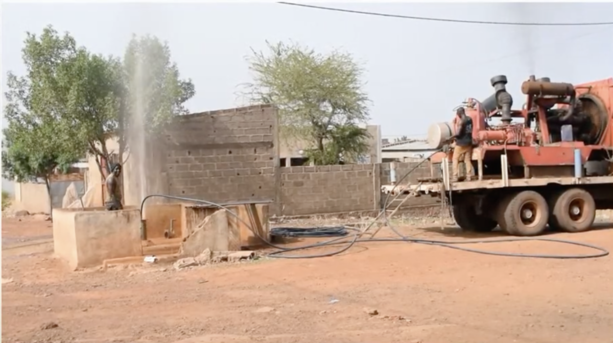 Water gushes from the ground while pipes run from the hole to a truck with large equipment in its bed.