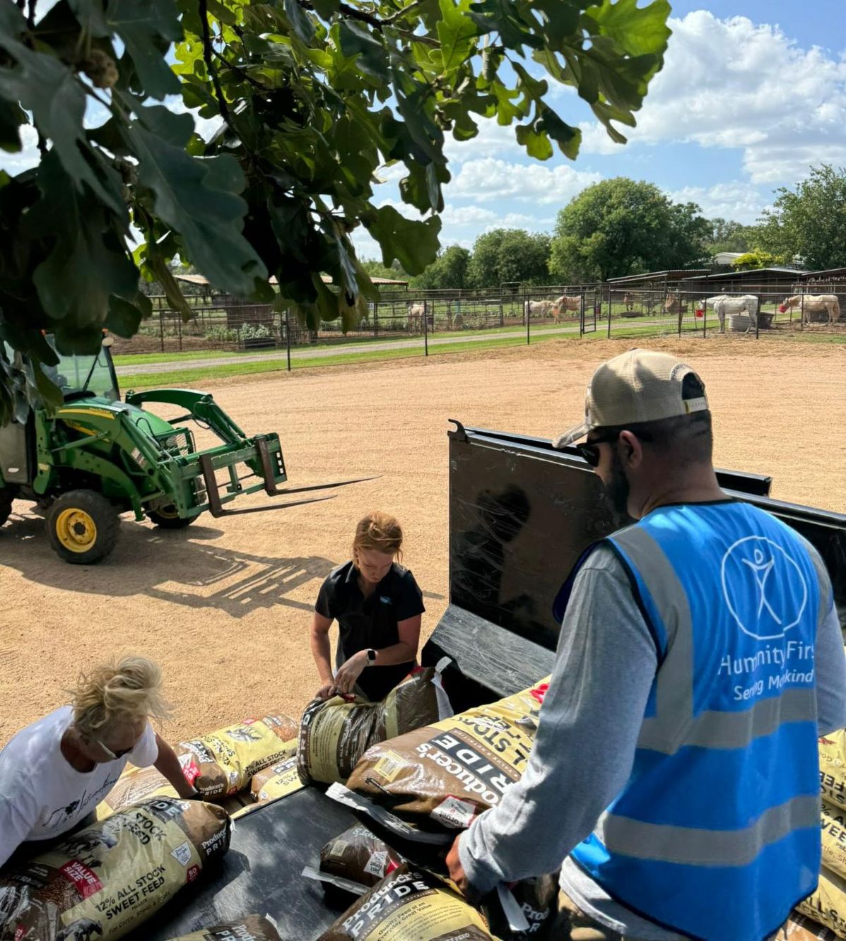A man wearing a blue humanity first vest helps unload heavy bags  of livestock feed from a truck on a ranch.