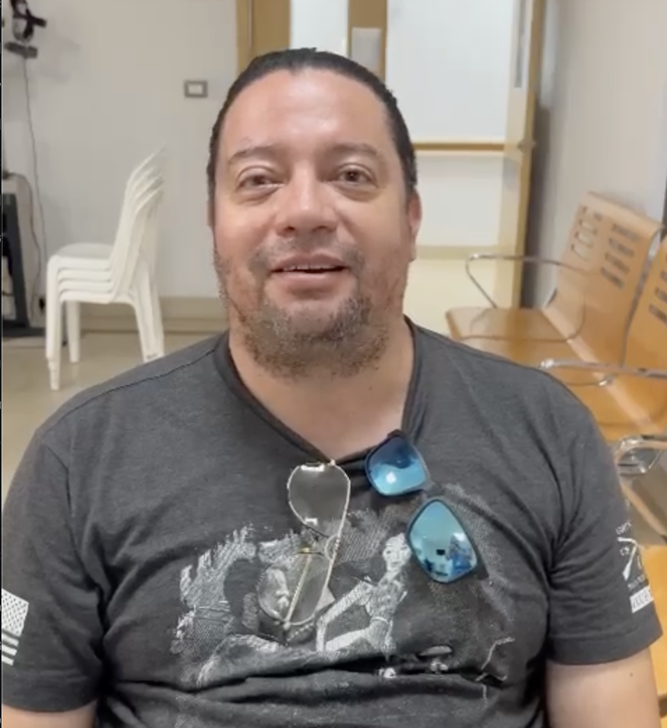 A man speaks to the camera from a hospital waiting room. He wears a t-shirt with glasses and sunglasses hanging from the collar.