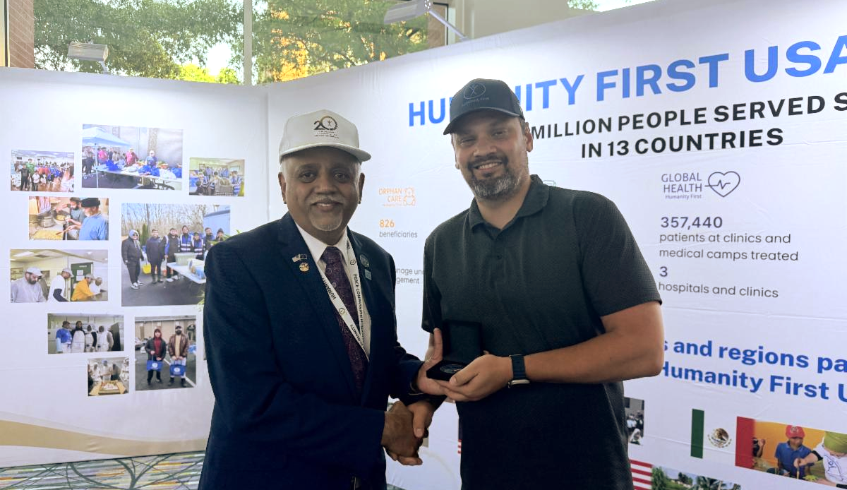 A man poses holding a Humanity First USA limited edition Challenge Coin and shaking the hand of our Executive Director.