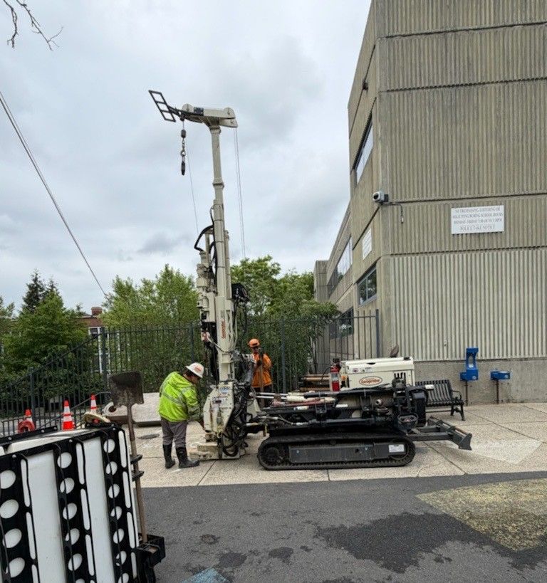 Two workers attend to a large drilling machine outside the former Winter Hill School Building at 115 Sycamore Street