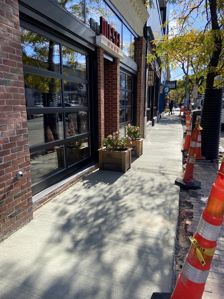 New sidewalks under construction in Davis Square outside Diesel Cafe; tall skinny construction cones are placed alongside the new concrete. 