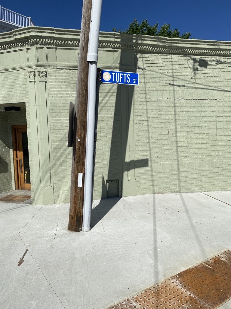 A new large sidewalk bumpout is seen in front of a green building. A street Tufts Street sign is on a poll in the sidewalk. 