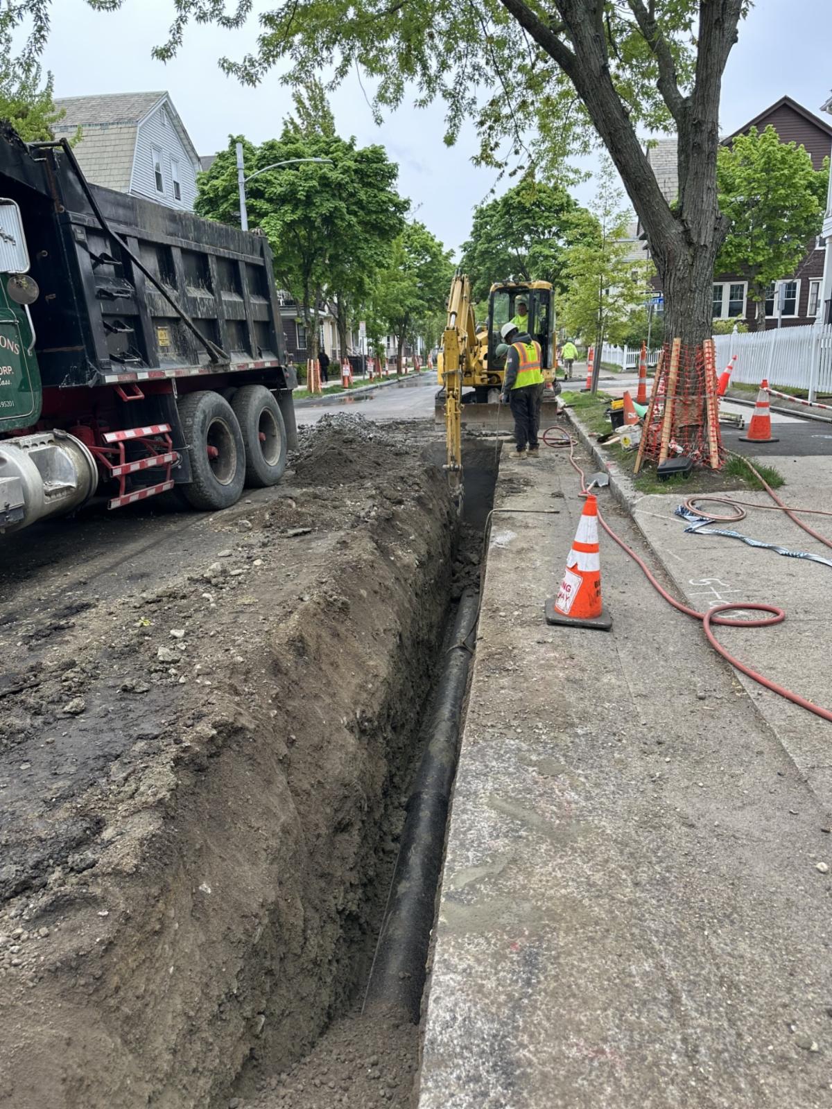 An open trench (dug up portion of the street) is seen with construction workers standing above it and a new pipe being put into the ground.