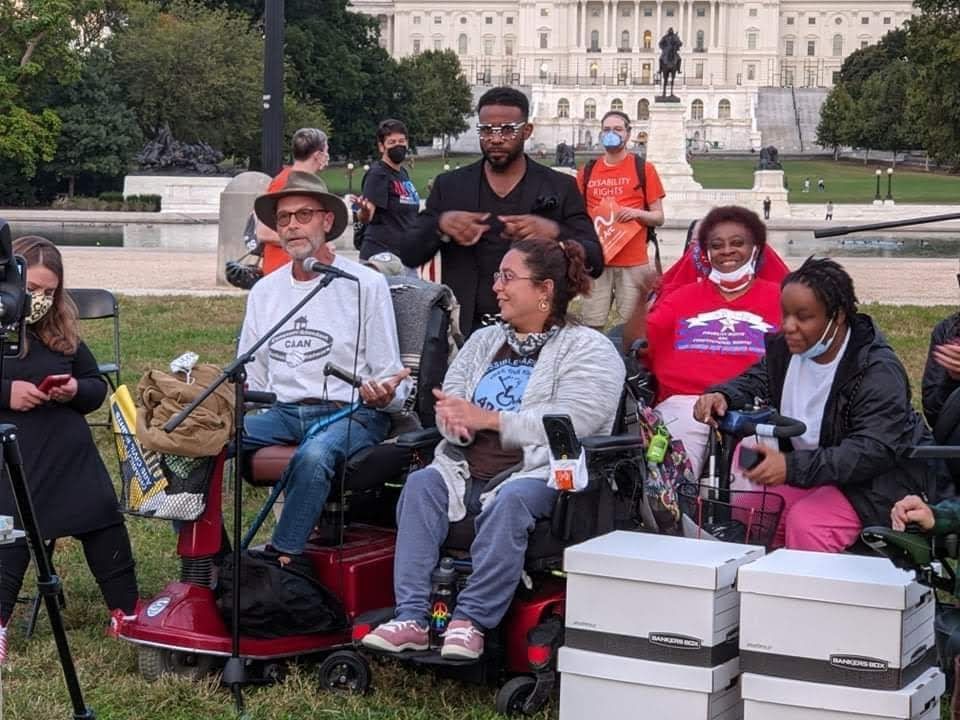 ADAPT members in mobility devices with an ASL interpreter behind them while speaking in front of the Capitol.