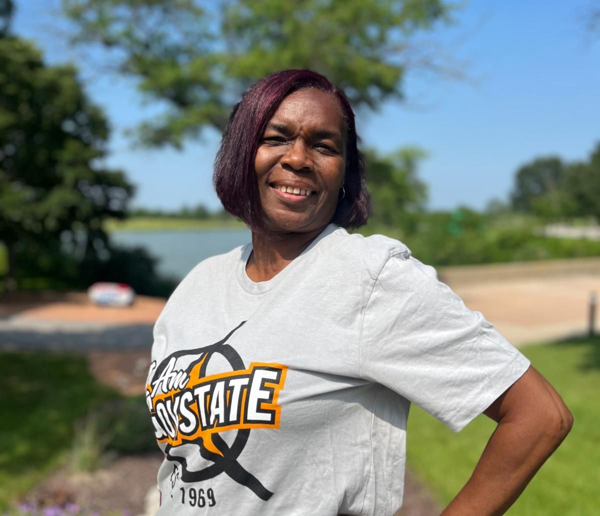 A smiling woman wearing a grey t-shirt that reads "I Am GovState" poses for a photo on the Lakeside Patio at Governors State University. Pictured is GovState alumna, Tammy Brown.