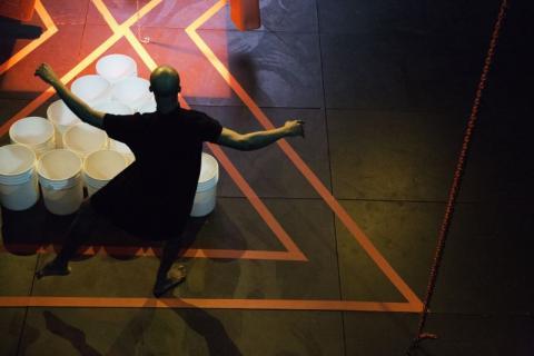 View from above of Arien Wilkerson dancing around large white buckets on on top of an orange and black tape design floor.