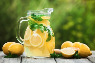 Four lemons are resting on a wood table beside a pitcher of lemonade with lemon slices and mint leaves.