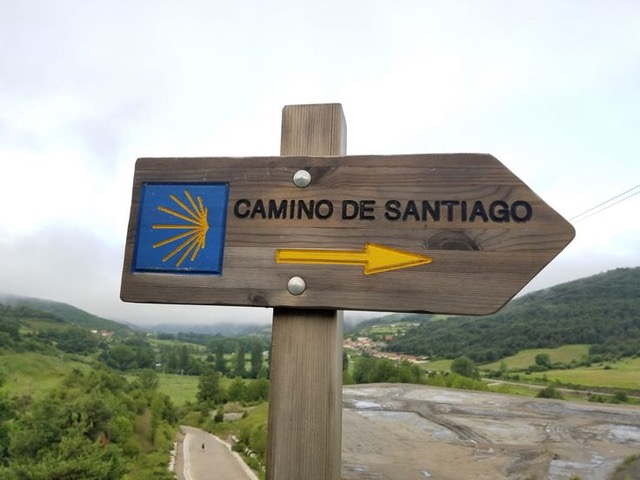 A wooden sign saying Camino de Santiago with a yellow arrow and a shell are in the foreground with green hills and the sky in the background.