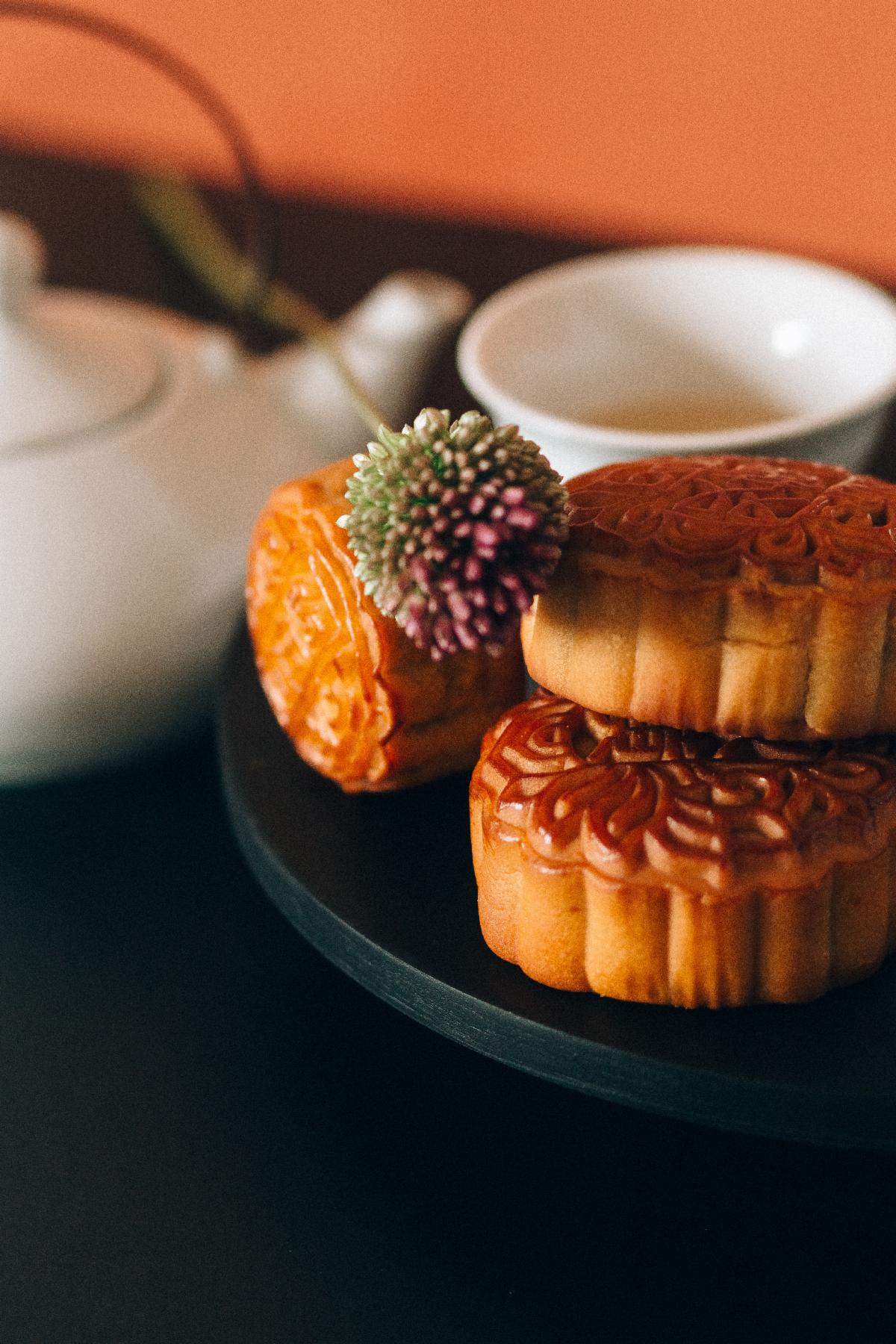 stack of 3 mooncakes with mum like flower on top, white teapot and porcelain cup with tea in the background. Orange backdrop