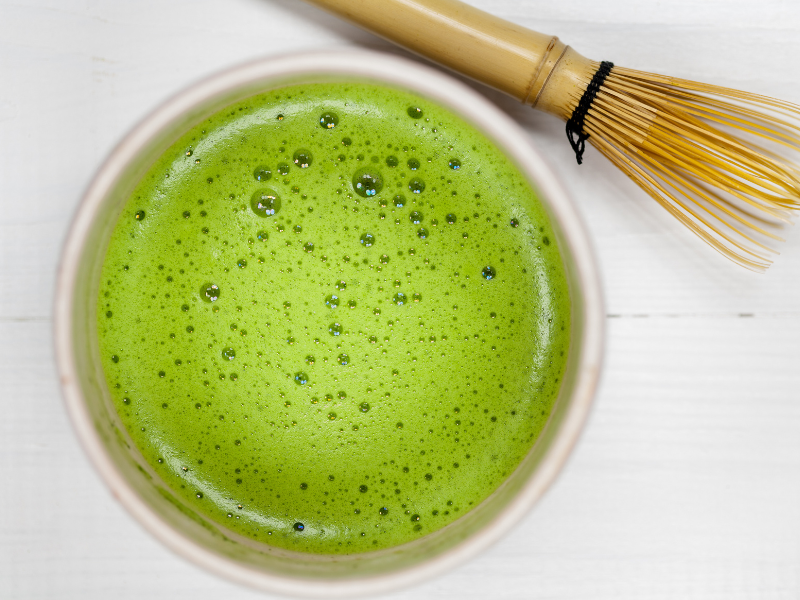 Very thick bright green frothy matcha bowl next to bamboo whisk on white table