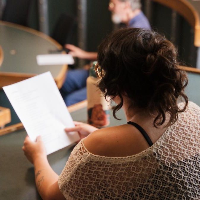 over the shoulder view of a woman in knitted top reading a white paper in classroom.