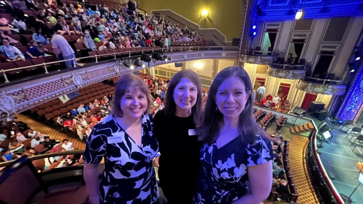 three women posed on balcony facing against a full theatre venue