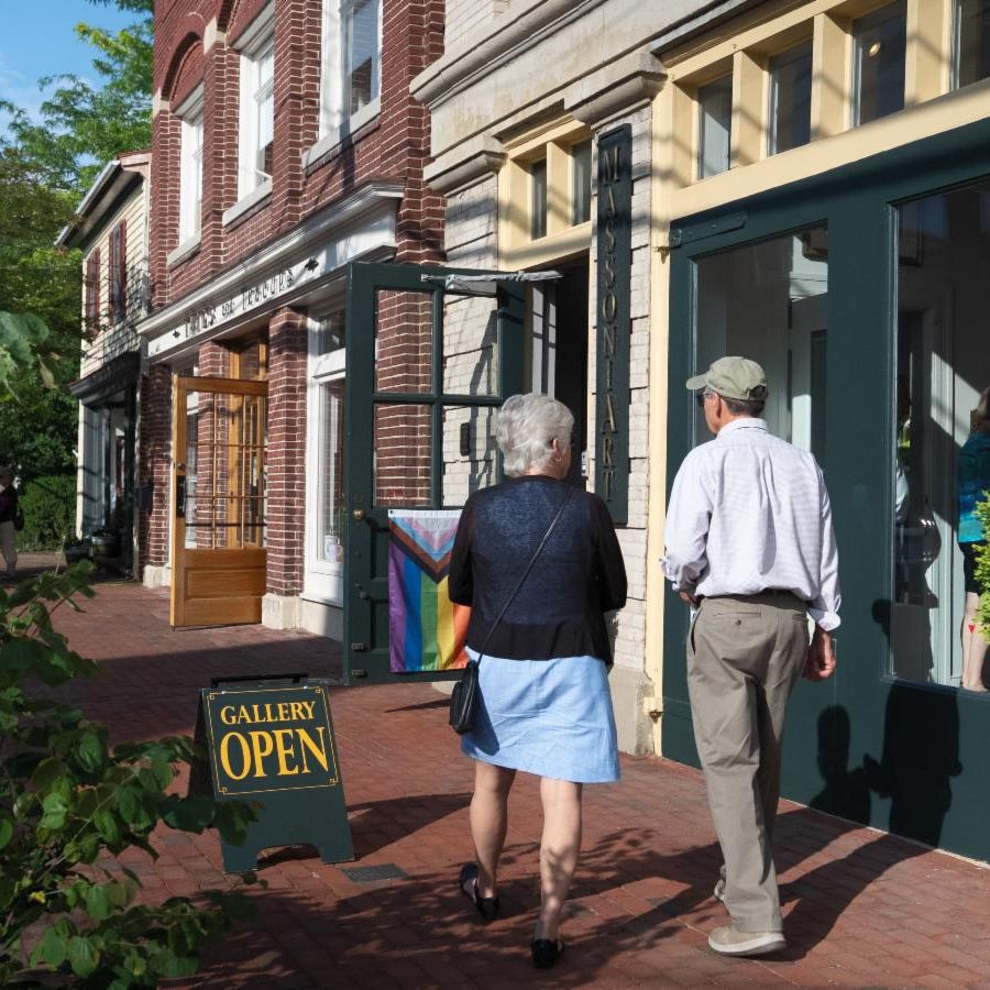 eldery couple walking by businesses in neighborhood passing a small green sandwich board sign that reads Gallery Open