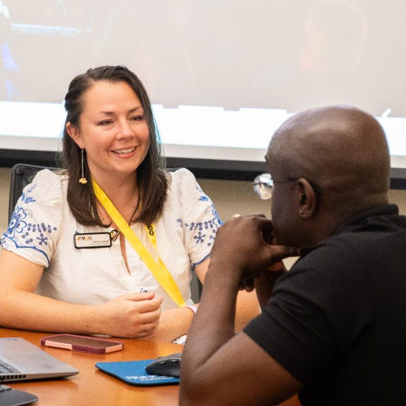 women in white dress speaking at a table with man in black shirt.
