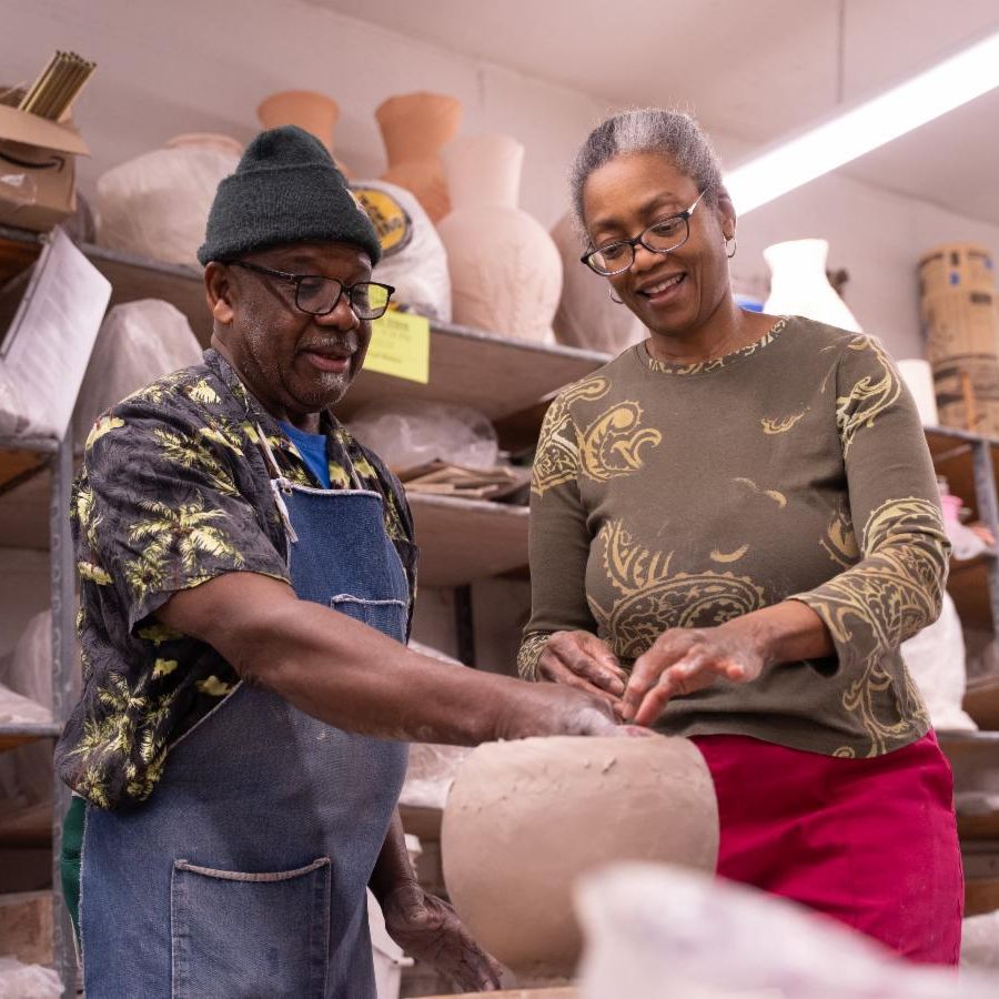 man and woman looking down at unfired ceramic pot surrounded by other unfired ceramics on industrial shelves.