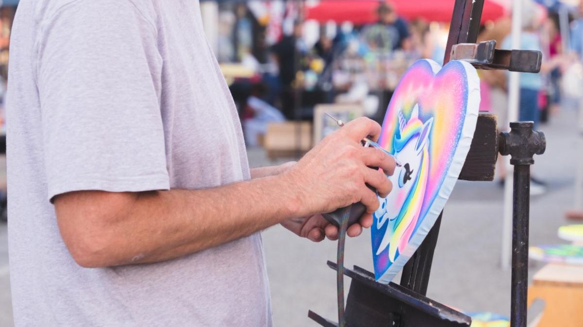 Male airbrush artist painting a rainbow unicorn on a heart shaped canvas on a black easel.