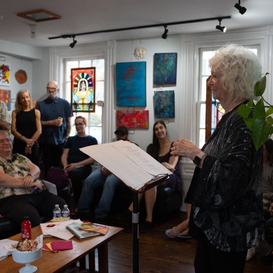 eldery women at podium speaking to audience in a blue room with two windows and paintings on wall. 