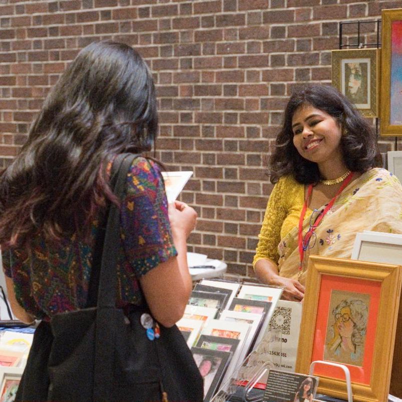 woman smiling at a customer standing in front of her vendor booth of handmade art. 