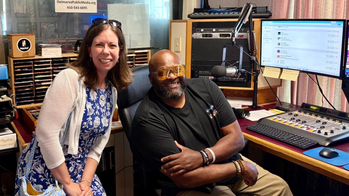 Seated man and standing woman smile for the camera in a radio station booth