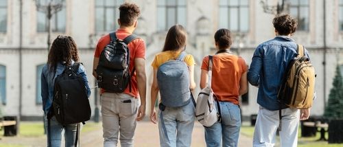Group of five students with backpacks walking at university campus together_ rear view of multiethnic young people going to classes_ diverse college friends spending time outdoors after lessons