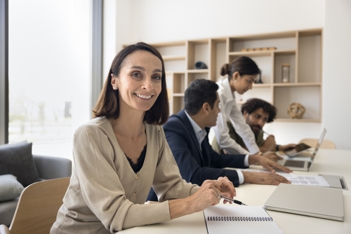 Happy mature Latin businesswoman_ project manager_ female team leader portrait. Positive business woman smiling at camera while with multiethnic colleagues working at laptop in background