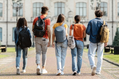 Group of five students with backpacks walking at university campus together_ rear view of multiethnic young people going to classes_ diverse college friends spending time outdoors after lessons