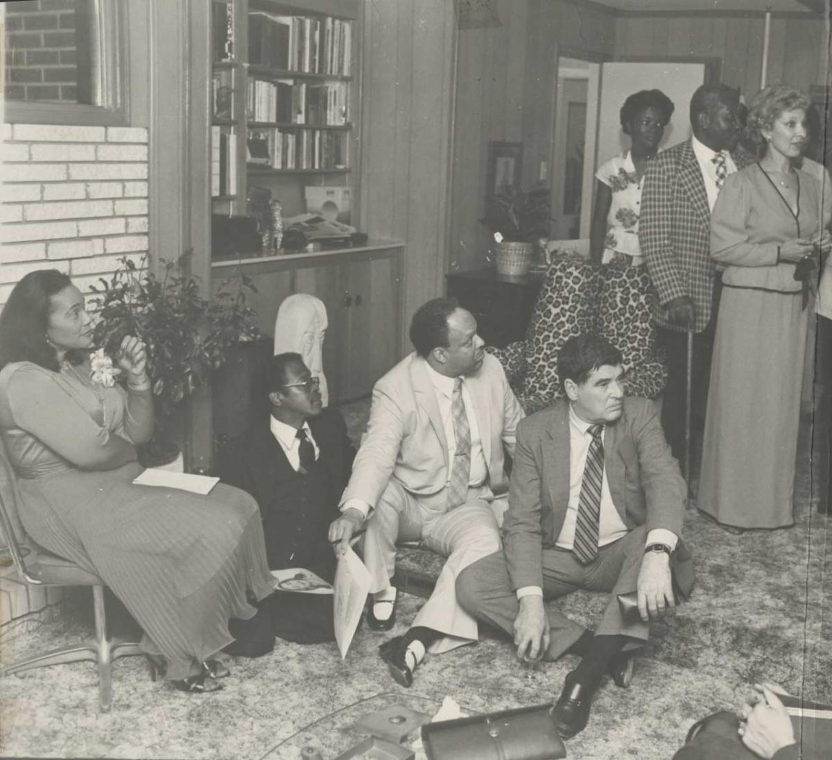 A black and white photo of several people sitting on the ground looking in one direction.