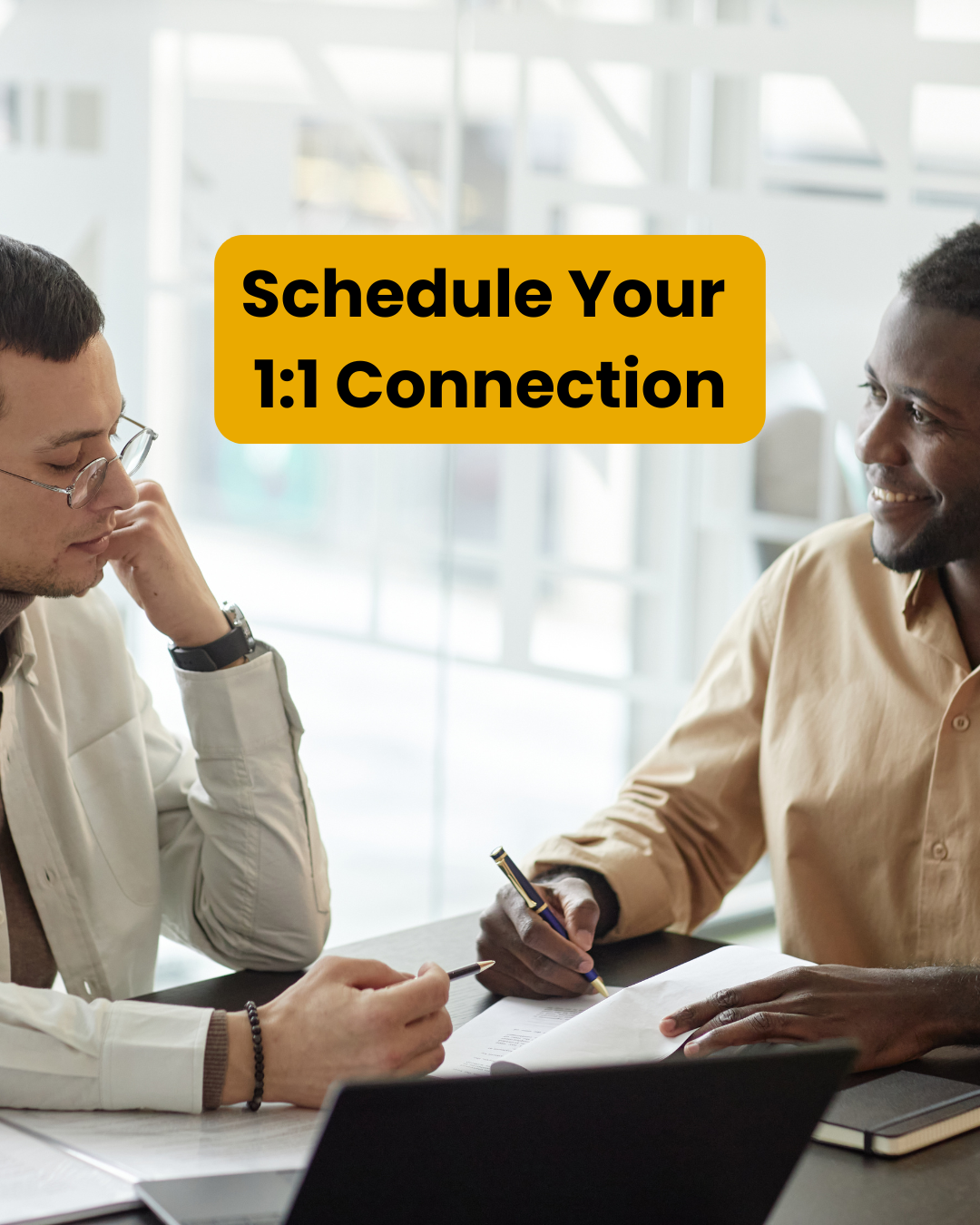 Two people sit across a table from each other in a bright office, one smiling while taking notes on paper and the other listening thoughtfully. Text reads: "Schedule Your 1:1 Connection.