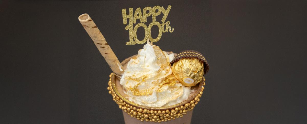Gold dessert drink topped with whipped cream, edible gold leaf, Ferrero Rocher candy, wafer roll, and âHappy 100thâ cake topper against a dark background
