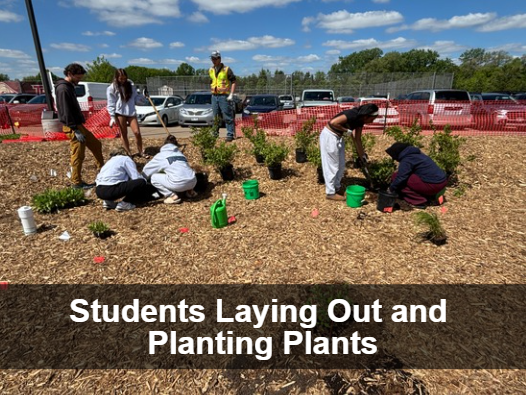 A group of people are planting bushes and small trees in a garden bed covered with wood chips. Green watering cans and buckets are visible, along with orange safety fencing in the background. Greyed out box near bottom with 'Students Laying Out and Planting Plants' in white text.