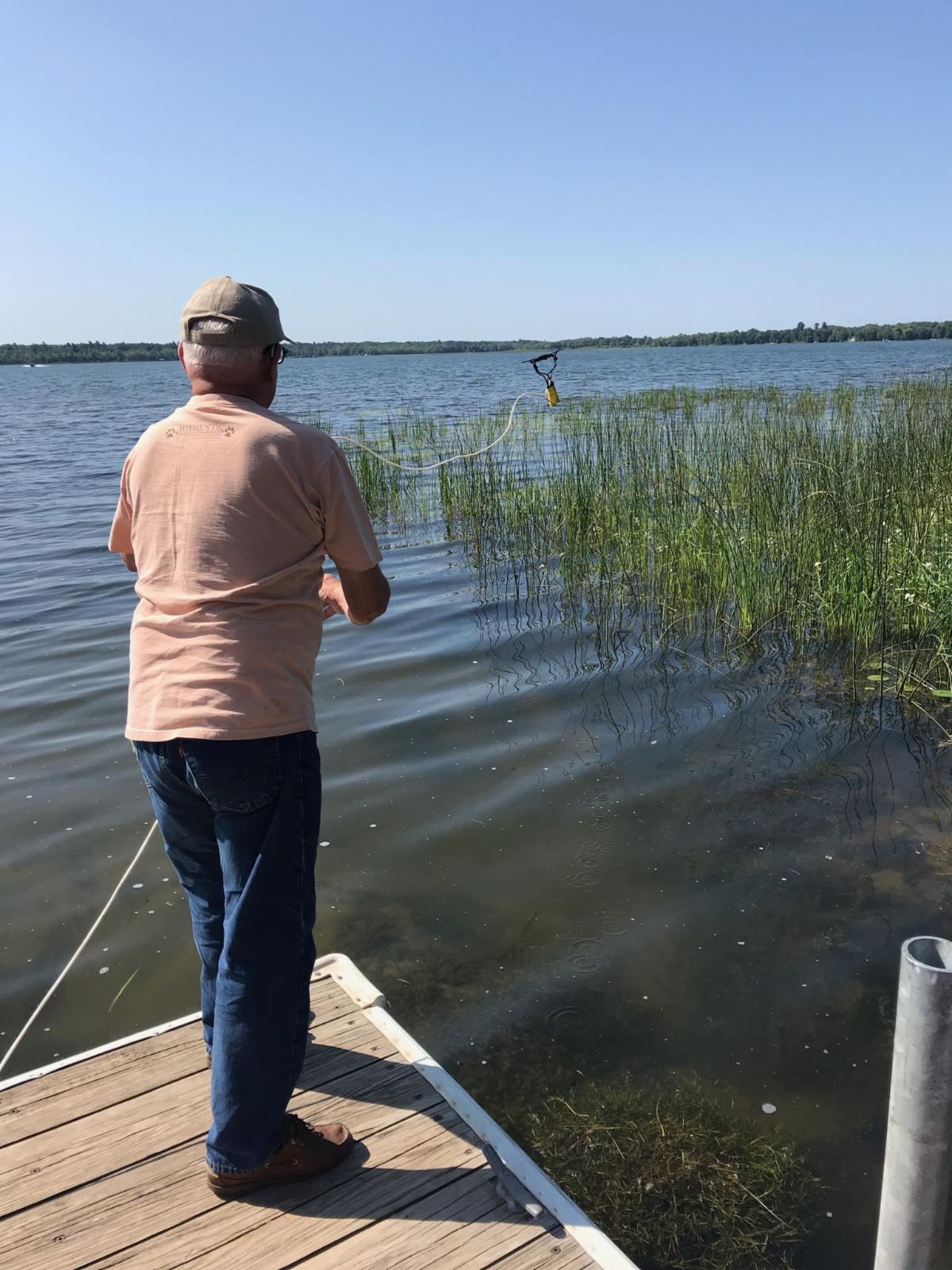 Man standing on edge of dock throwing rake towards emergent vegetation in water.