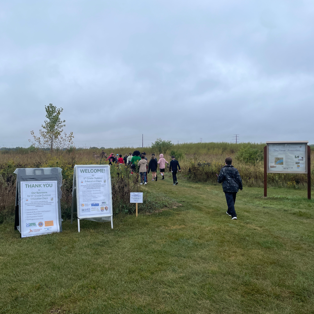 People walking along a grassy path at the prairie entrance, passing the “Welcome to Outdoor Ed Days” and “Thank You Sponsors” signs under an overcast sky.