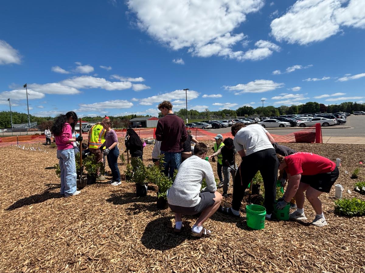 A group of people planting shrubs and trees in a mulched area under a partly cloudy sky.