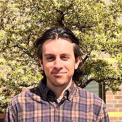A person wearing a plaid shirt stands in front of a tree and a brick building.