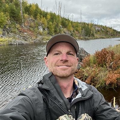 A person wearing a fishing vest and cap smiles at the camera with a lake and autumn foliage in the background.