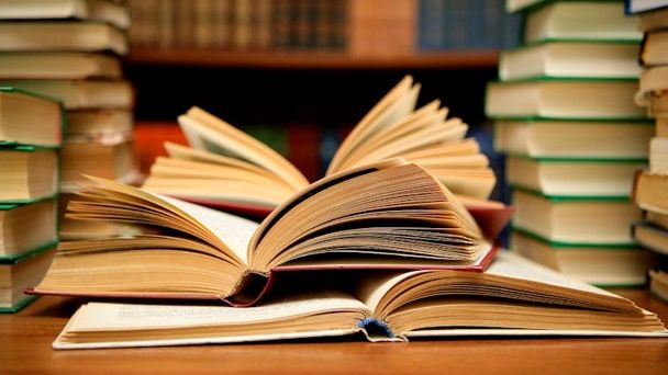 Books lay open on a smooth wooden table. Piles of closed books surround them, with more on the shelf behind them.