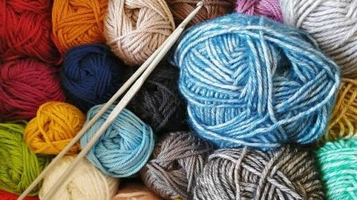 Colorful balls of blue, red, pink, yellow, gray, and green yarn fill the image with white knitting needles laying on top of them.
