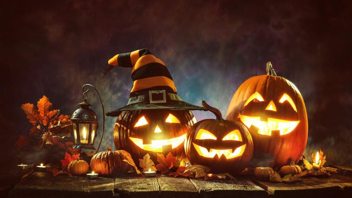 Jack o Lanterns glow in the night on a wooden table next to a lantern, candles, and fall leaves. A black and orange witch hat sits on the Jaco o Lantern on the left.
