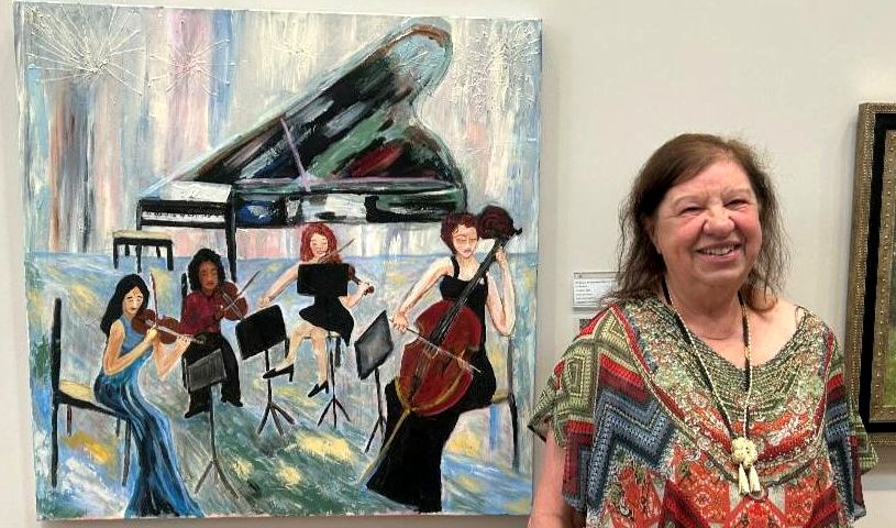Lori Packlick (shoulder-length brown hair, smiling, caucasian, wearing a green, blue, and red patterned blouse with black pants) stands next to her painting of a 4-woman orchestra playing in front of a grand piano.